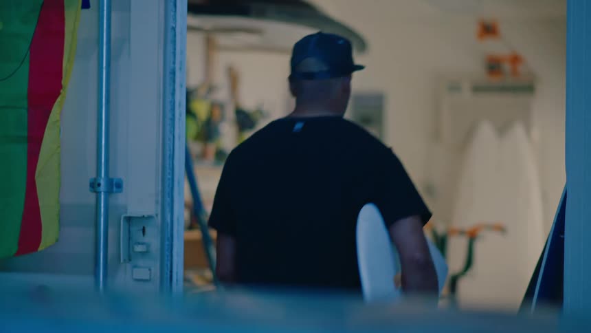 Man in cap picking up a white surfboard and placing it inside an indoor room