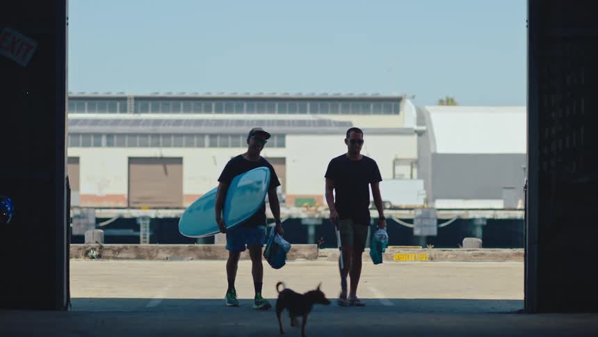 Two friends walking side by side each carrying a surfboard under one arm entering a warehouse