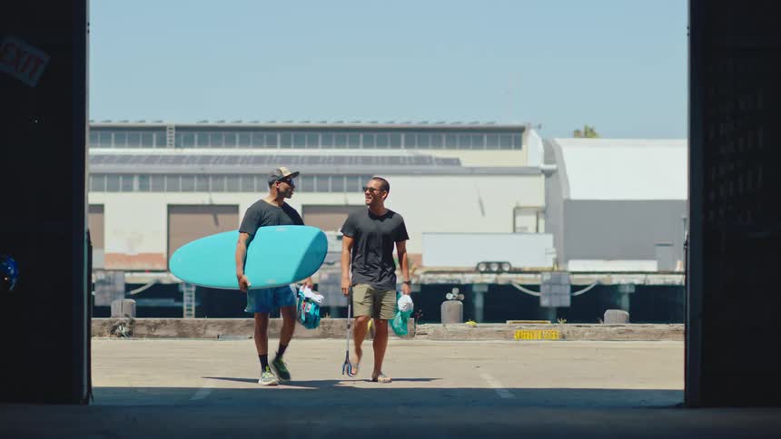 Two people walking into a place and one of them is holding a big blue surfboard