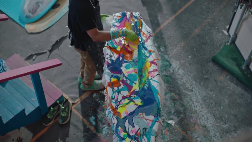 A man paints a surfboard various colors inside a workshop