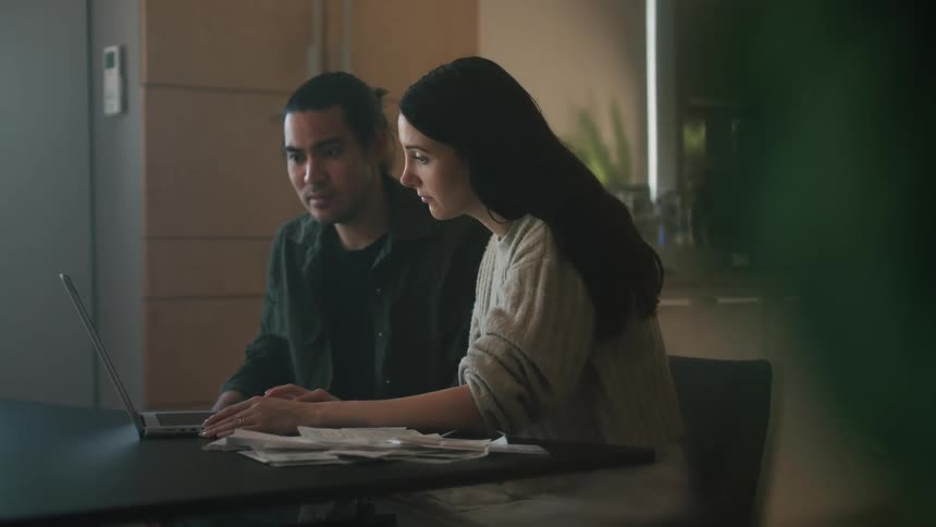 A couple sits at a table near the kitchen talking while focusing on a laptop