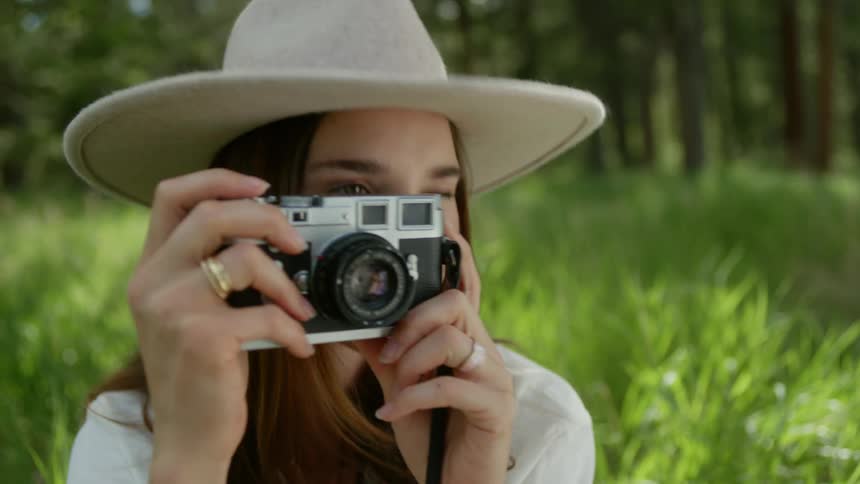 A woman holding a vintage camera to her face capturing a photograph in a lush green outdoor setting