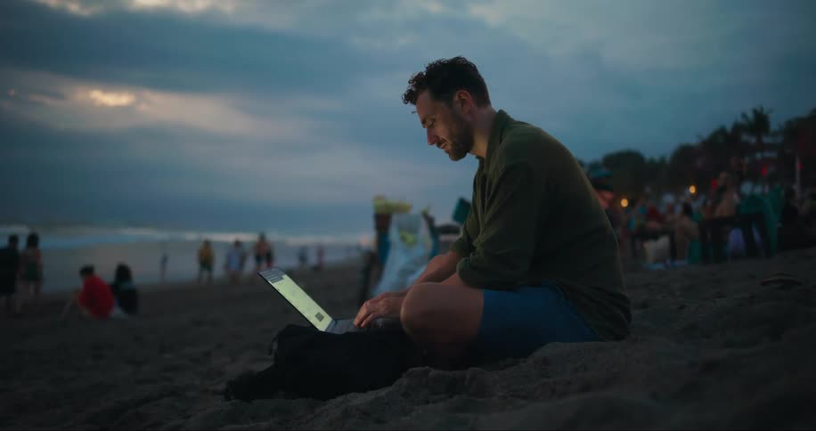 Man sits on a busy beach after dusk and types on a laptop