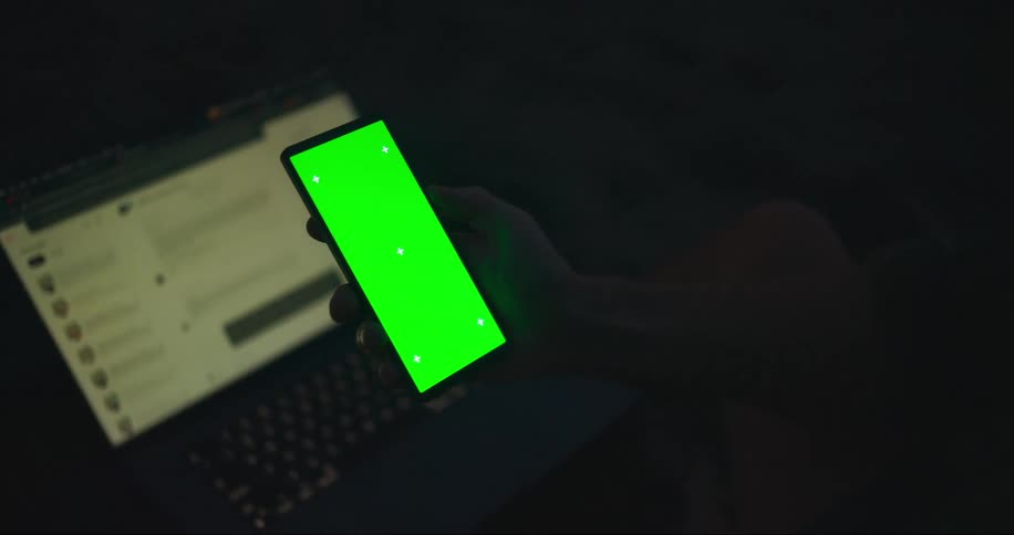 A hand holds a smartphone with a green screen in front of an open laptop in a dimly lit environment