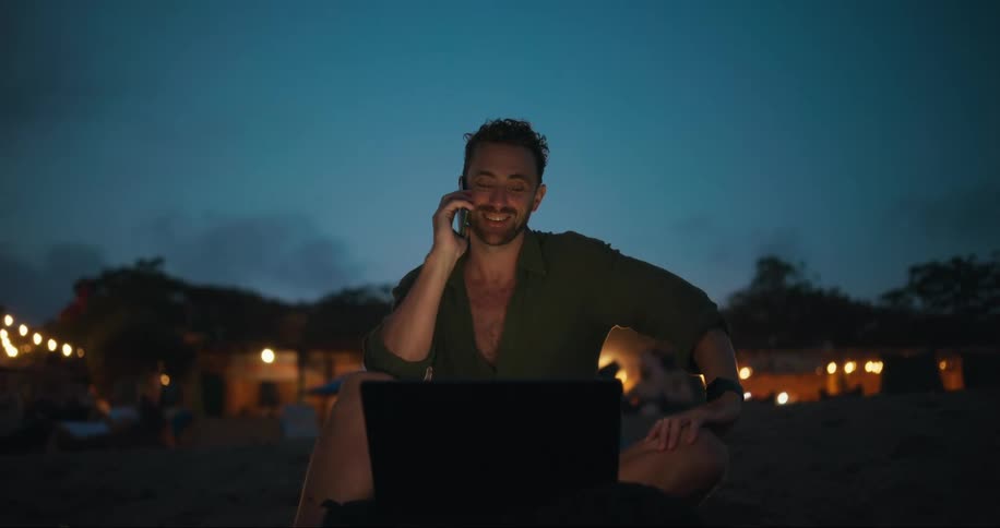 Man sitting on the sand talking on his cell phone and with a laptop in front of him in a nighttime environment with houses lit up in the background