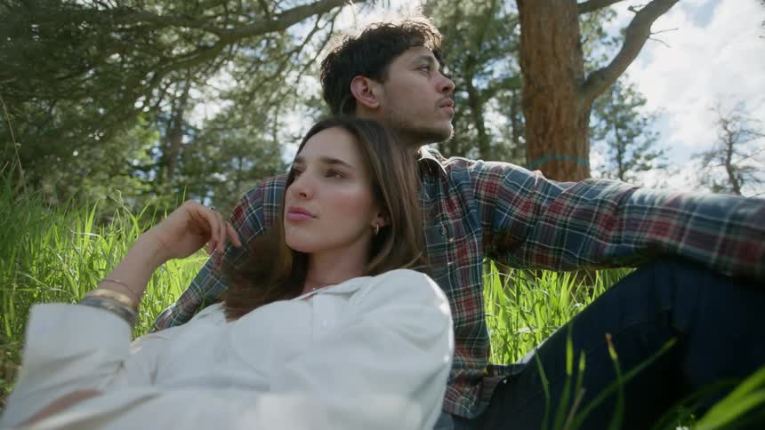 Woman in white clothing sitting on the chest of a man wearing a plaid shirt in a natural environment with trees around