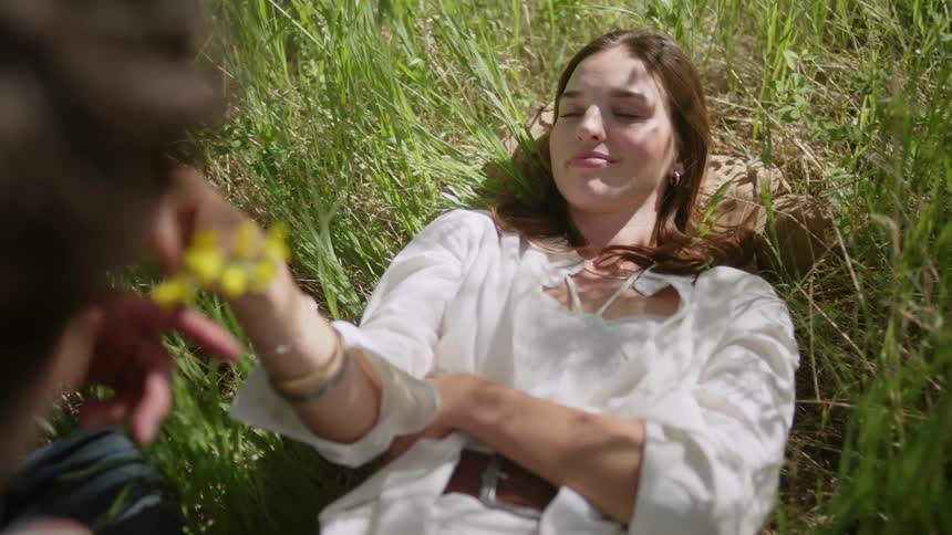 A woman lying in a grassy field smiling and reaching out to someone while holding a flower