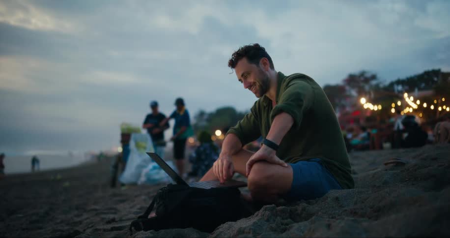A man smiles while using his laptop on the beach
