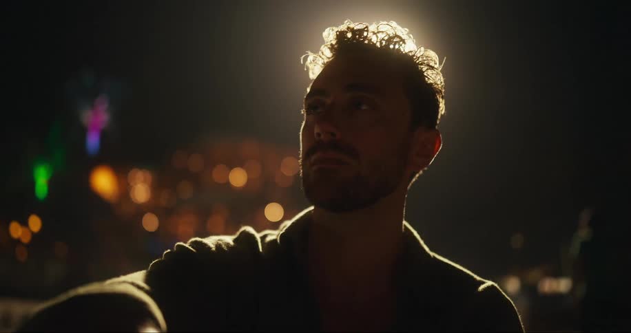 A white man with a beard and a halo of light around his head sits near an outdoor gathering