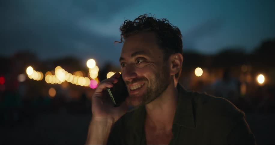 A closeup of a young mans face as he talks on a smartphone outdoors with blurred lights in the background at dusk