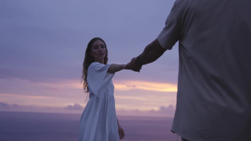 Woman in white dress holding hands with a man in a seaside setting with the sky in shades of orange and purple
