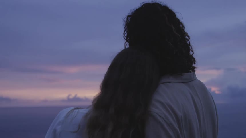 Back view of a man and a woman embracing each other affectionately while admiring a view of the sea and a sky with shades of purple