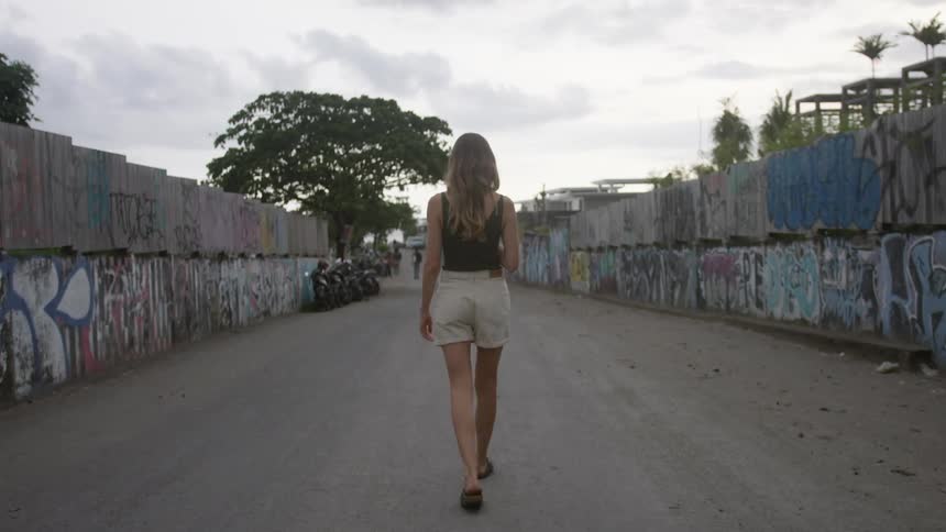 View of a woman from behind wearing beige shorts and a black sleeveless shirt walking in a room with graffitied walls
