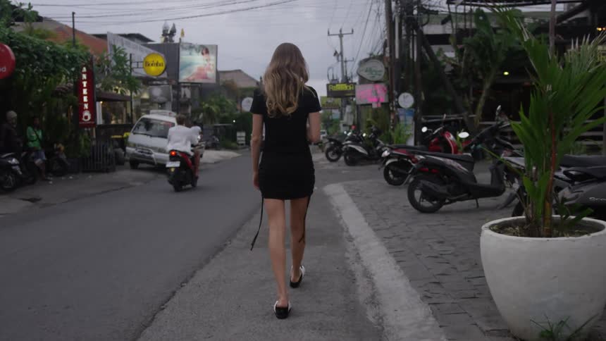 A white woman in a black dress strolls down a road in a city as motorcyclists pass by
