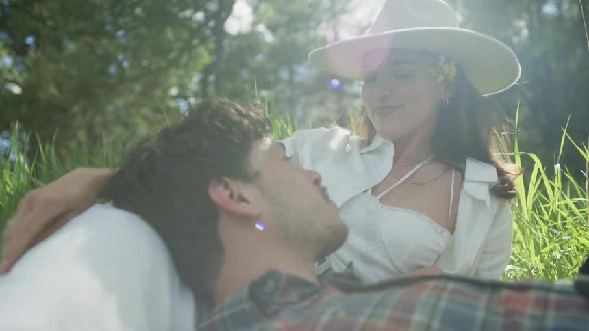 A man places a flower behind his womans ear while they recline on grass in a forest