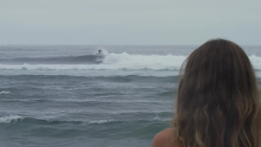The back of a womans head as she watches people surfing in the ocean