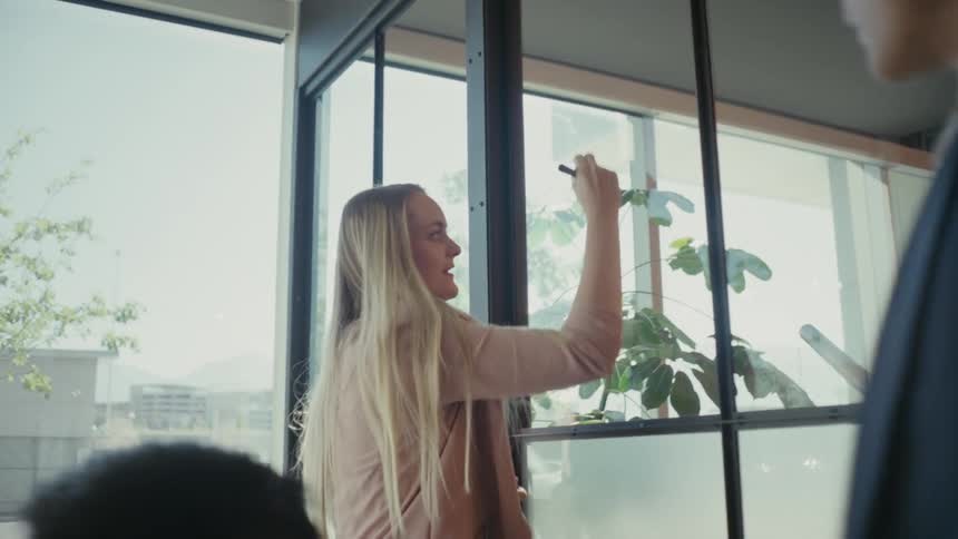 A woman in a pink blazer using a marker to write on the window of a meeting room