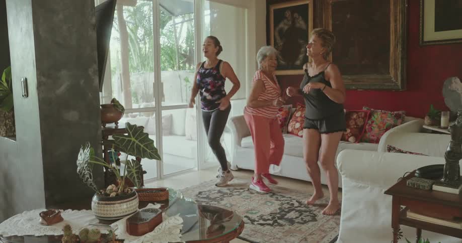 Older female friends exercise together in the living room at home on a summer day
