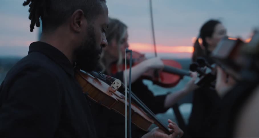 A group playing violins against a dusky sky with a city skyline behind them