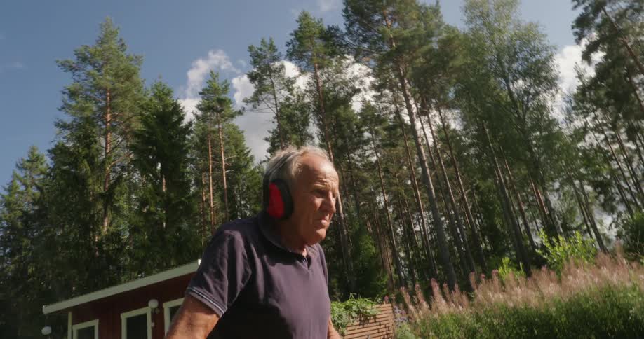 An old man wearing headphones walks outside near a forest