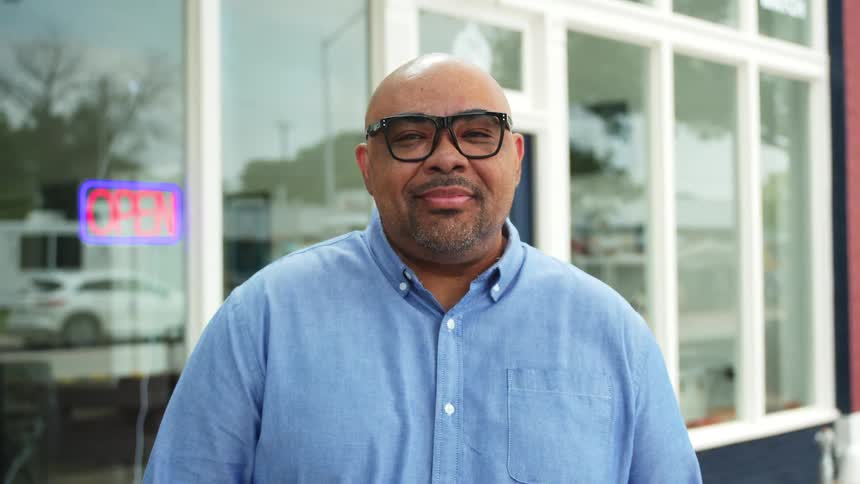 The face of a bald man with reading glasses who is looking to his side and then looking forward and is wearing a partial smile