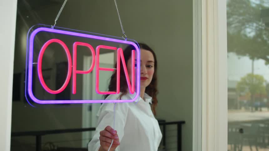 A woman stands behind a glass door turning off a neon sign reading open in bright pink and blue colors