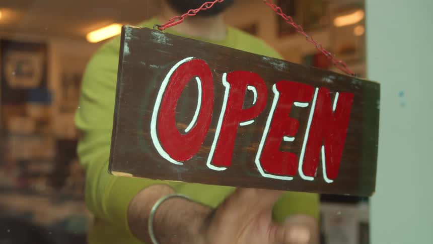 A bearded man wearing a blue turban turns around a red sign with the word open