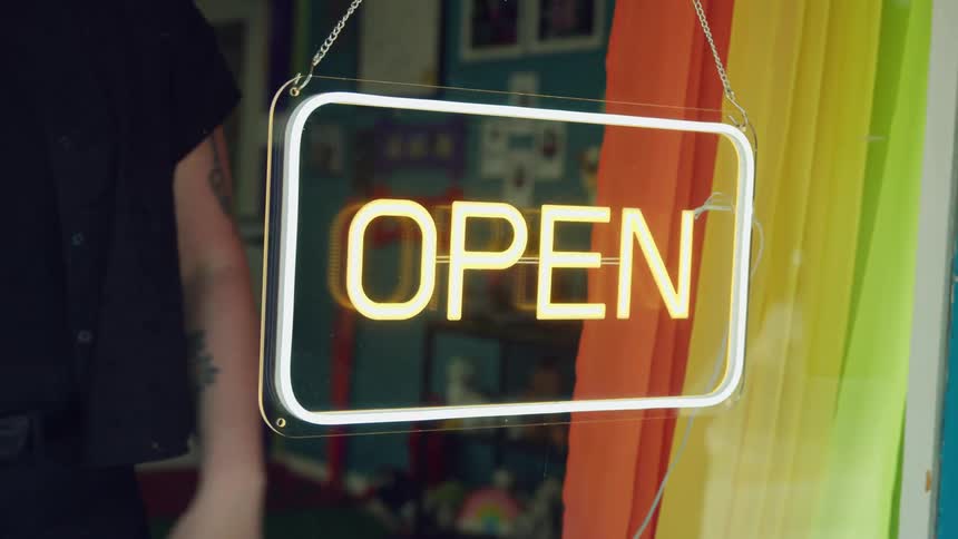 A woman stands behind a glass door turning on a neon sign reading open in bright yellow and white colors