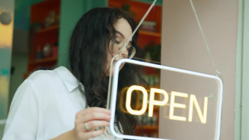 Young woman turning on a sign with the word open that is on the window of a door of a commercial establishment