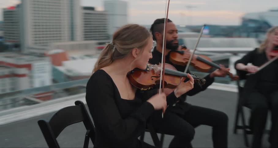 A string quartet is performing on a rooftop with musicians playing violins and a cello and a cityscape in the background