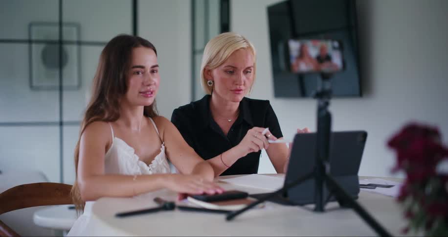 Two women sitting at a table being recorded by an iphone with a tablet in front of them