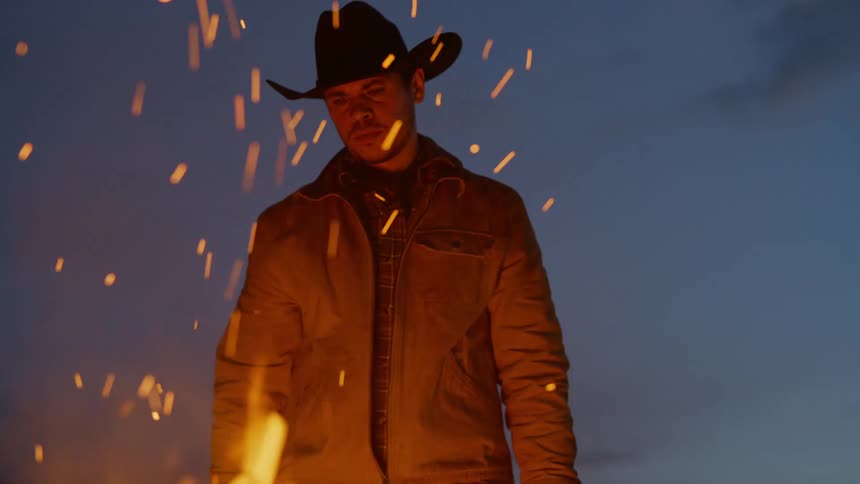 Cowboy in hat in front of a campfire at night putting firewood
