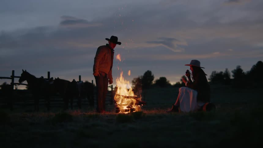 Man putting firewood on a fire in a field and a woman sitting next to him photographing the scene on a dark late afternoon