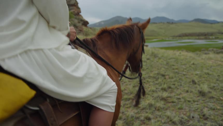 A white woman rides a horse near a body of water on a grassy plain