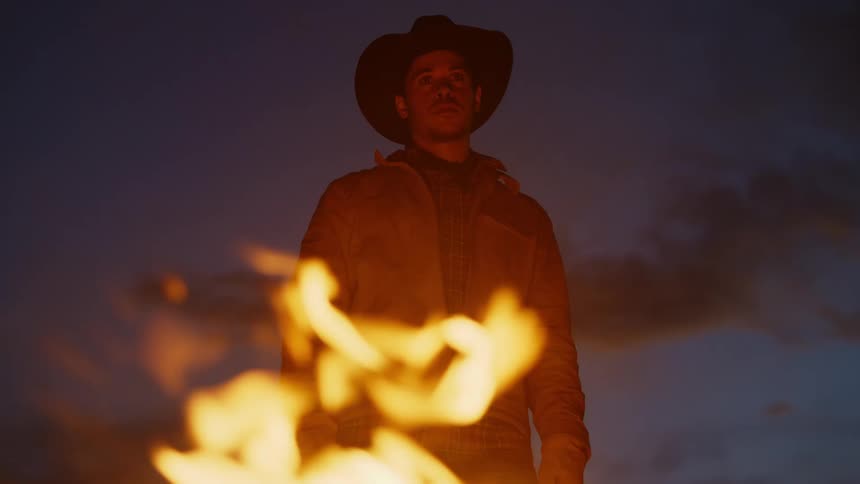 Man putting on a hat outdoors in front of a campfire at night