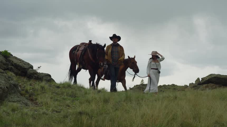 Man and woman in hats walking with two horses in a hilly setting on a cloudy day