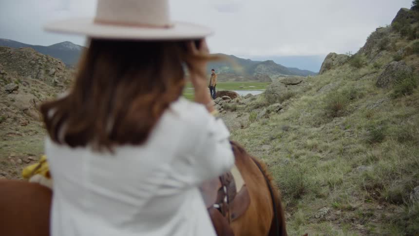 A woman in a cowboy hat stands next to a horse and she picks up a camera to take a picture of a man who is standing in the distance and waiting