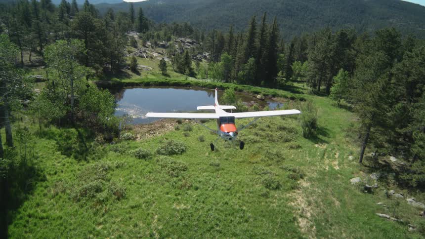 A small airplane is flying in slow motion over a lush green landscape with a pond and a mountain range in the background