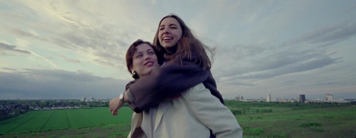 Two young women are joyfully interacting outdoors with one giving the other a piggyback ride through a green field with a city skyline in the background