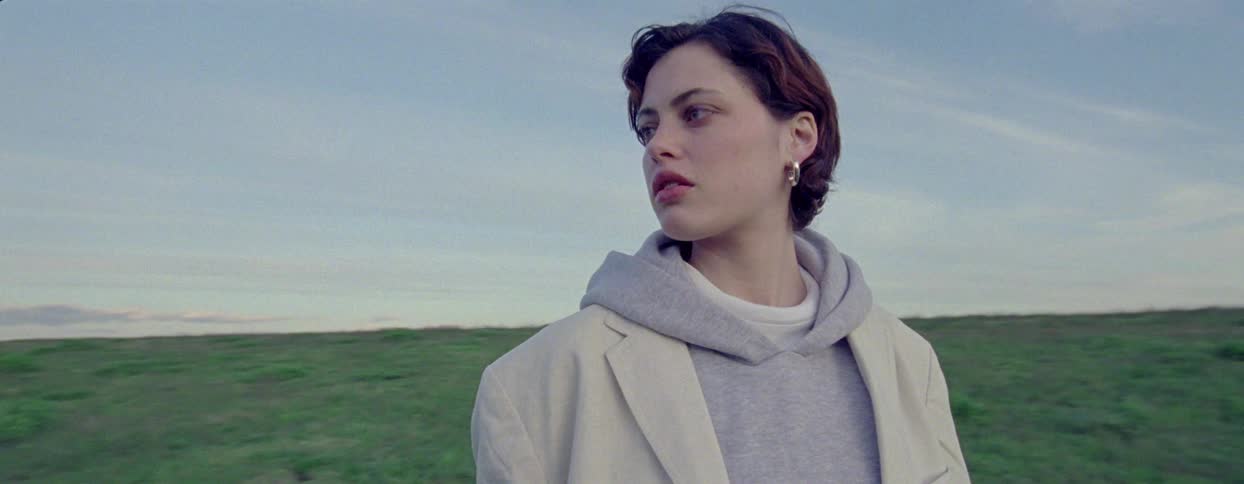 A woman walks in a grassy field while looking around somewhat nervously