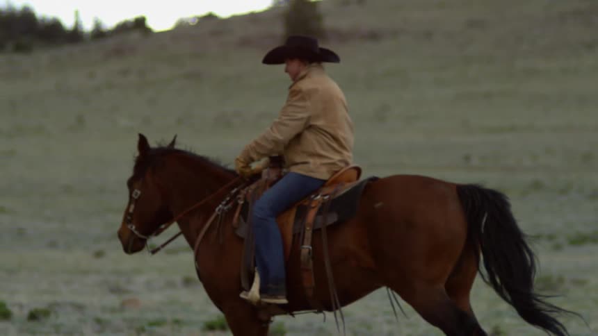 A person riding a horse across a grassy landscape in slow motion wearing a cowboy hat and a light colored jacket