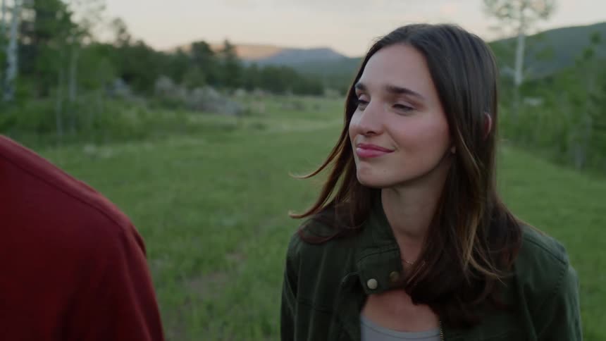 A young woman has a small smile on her face as she is talking to a man in a red shirt who is walking beside her and they are both walking in a grass field