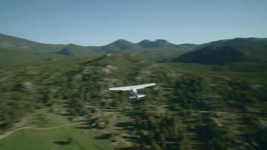 Small plane flies over a rural homestead surrounded by forested hills and mountains on a sunny day