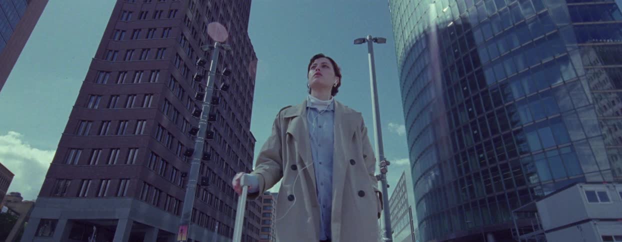 A woman wearing a coat and pushing a piece of luggage looks around as she walks in a city