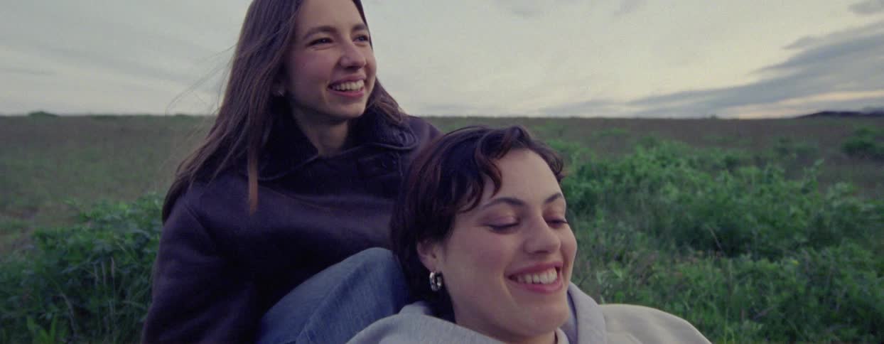 Two women are sitting in a grassy field talking and gazing at each other with smiles