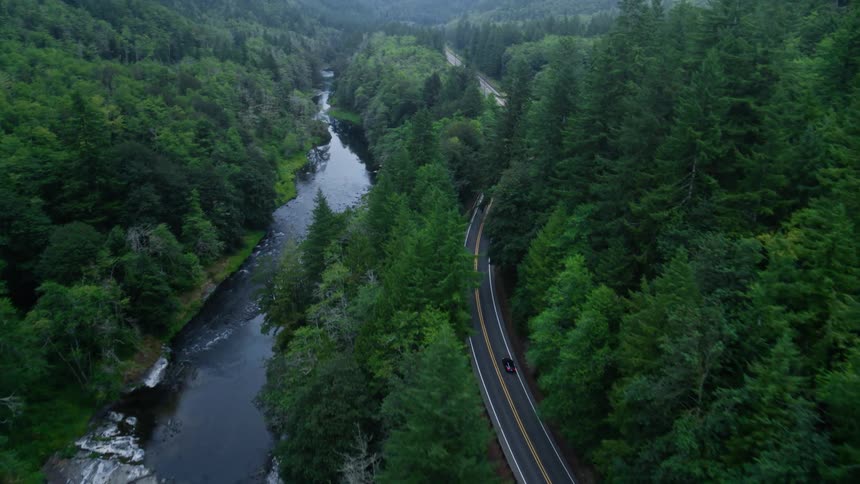 An aerial view of a car driving along a road through a dense forest in the mountains near a river