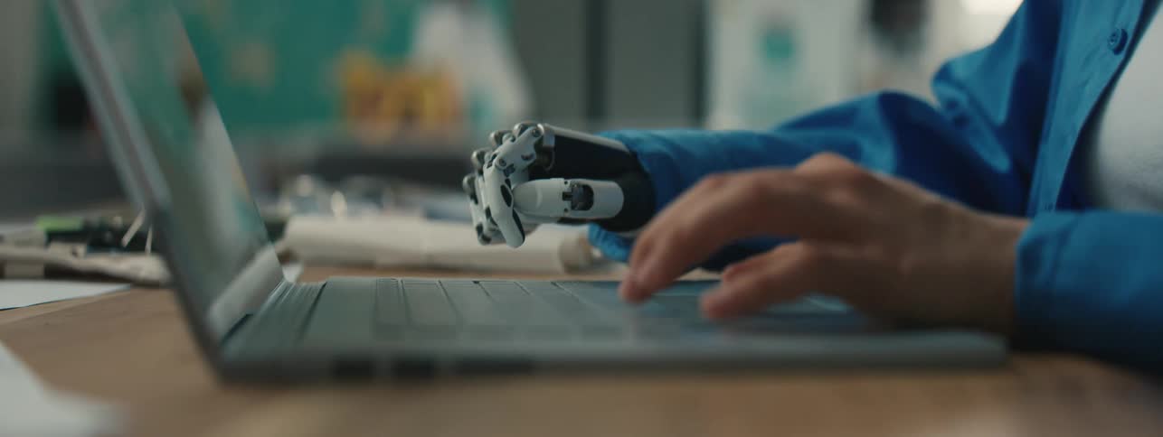 Woman with one robotic hand and one human hand typing on a laptop