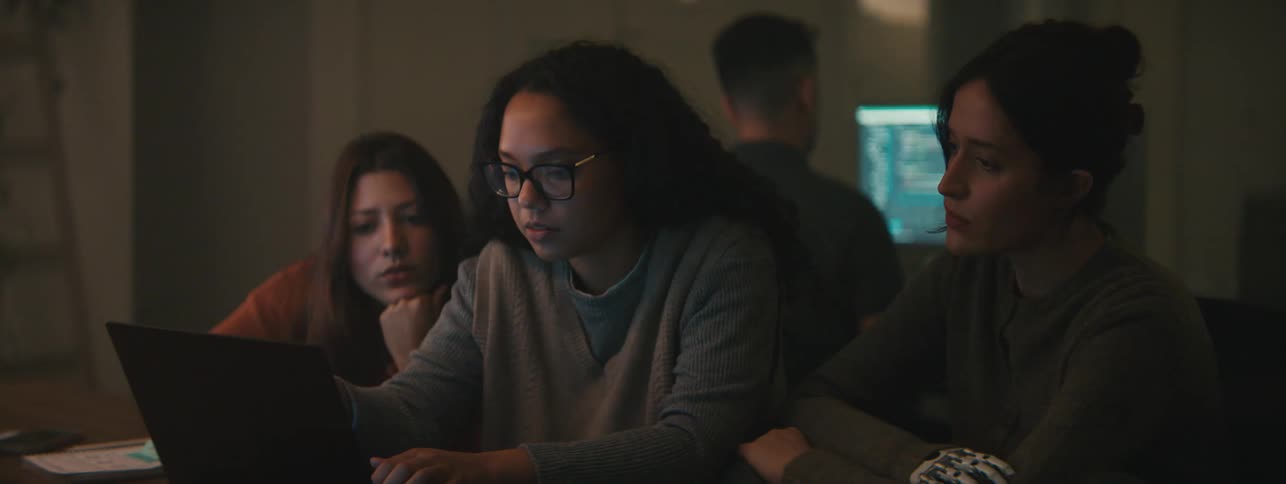 Three individuals collaborating on a laptop in a dimly lit environment with a person in the background working on another screen