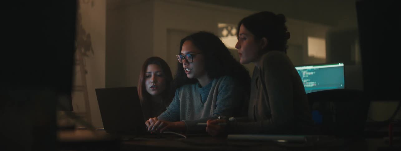 Three women collaborating over a laptop in a dimly lit workspace