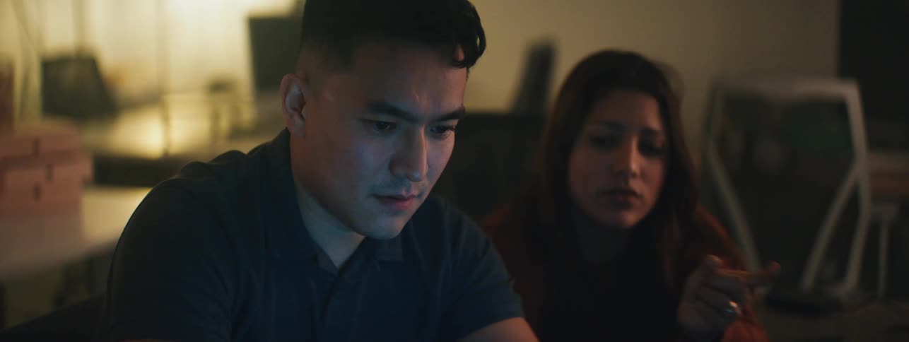 Light from a computer screen illuminates the face of a man collaborating with a female coworker in an office